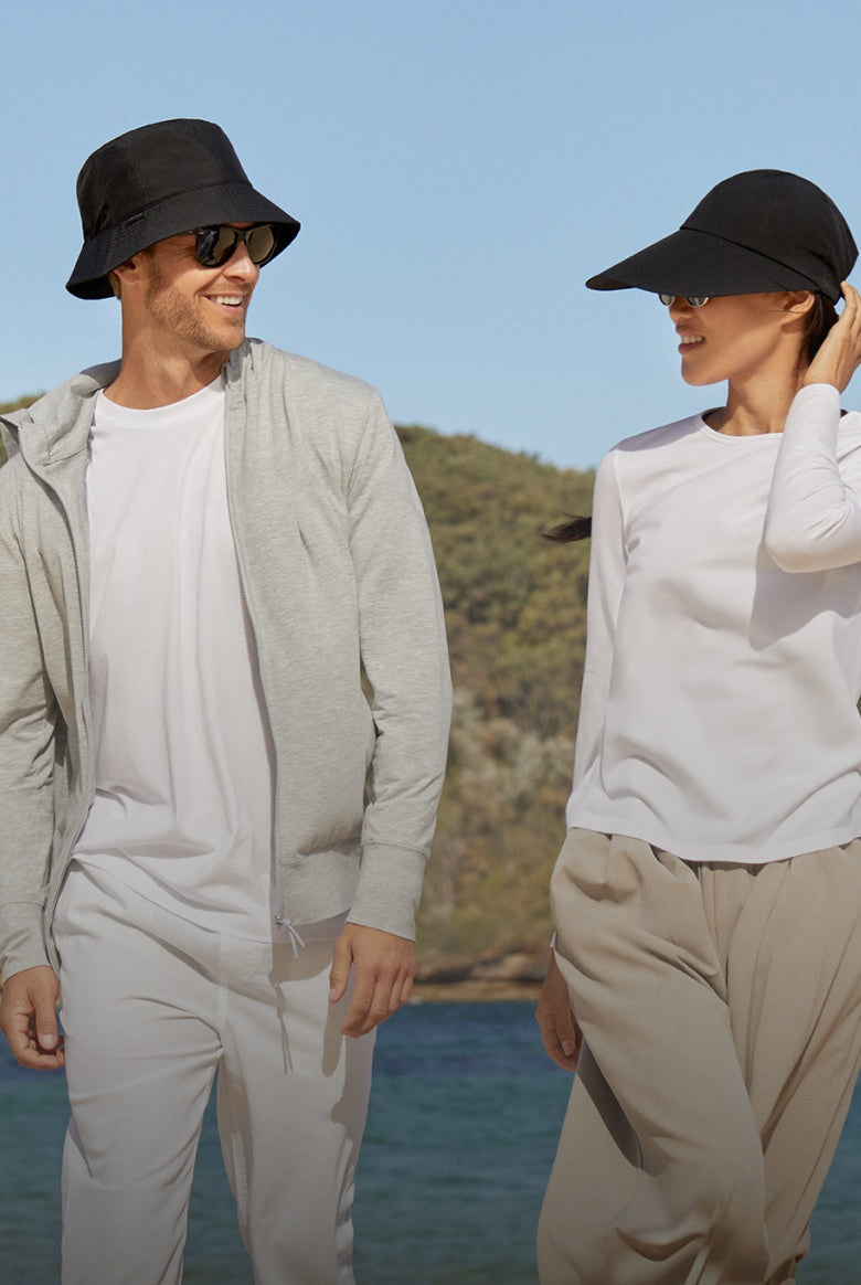 Two people wearing black hats and sunglasses standing by a body of water with mountains in the background.