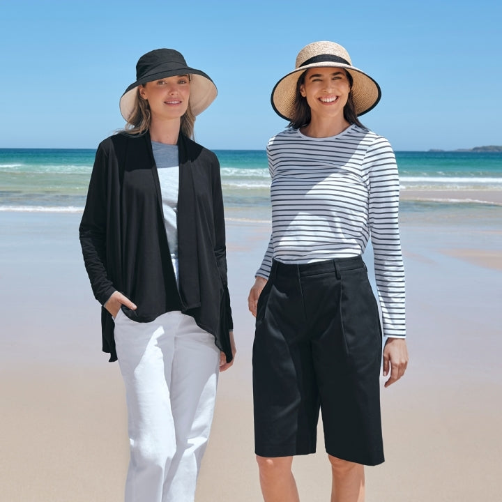 Two women standing on a beach wearing hats and casual clothing.
