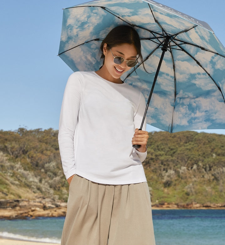 Woman holding a cloud-patterned Solbari sun umbrella by a beach.