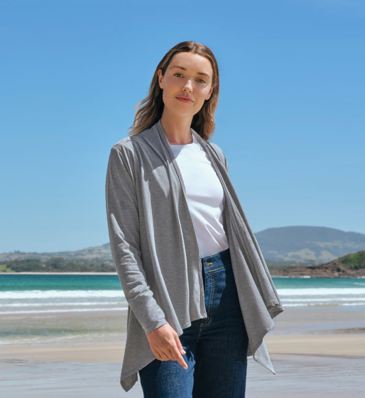 Woman wearing a Solbari UPF 50+ grey cardigan over a white shirt with a beach and blue sky in the background.