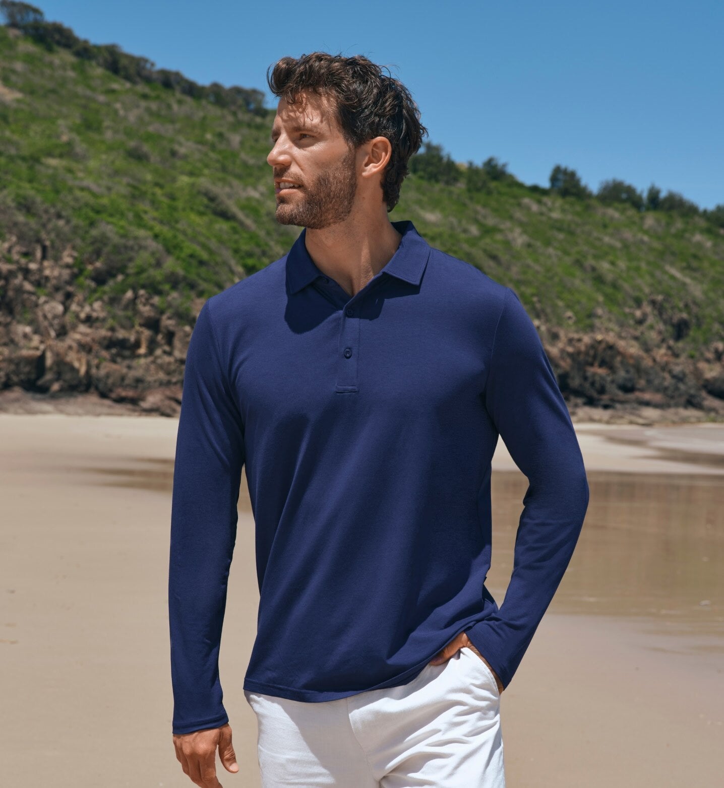 Man wearing a navy long-sleeve polo shirt and white pants standing on a beach.